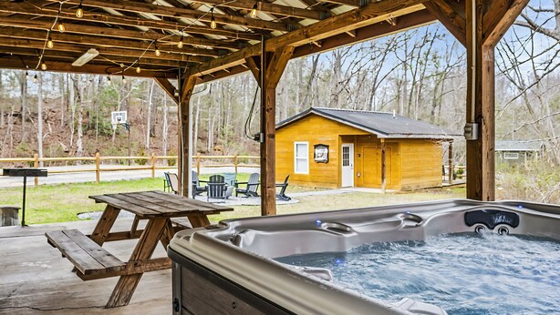 Un quiosco de madera con un jacuzzi, mesa de picnic y vistas a una cabaña al fondo.