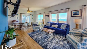 Ein Wohnzimmer mit blauem Sofa, einem Couchtisch, einem Essbereich mit Stühlen, einem an der Wand montierten Fernseher und Meerblick durch große Fenster.
