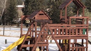 Une structure de jeux en bois, dotée d’un toboggan et de balançoires, installée dans un espace enneigé, avec des arbres en arrière-plan.