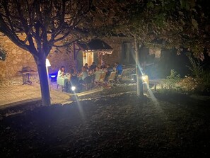 An outdoor dining area with tables and chairs under a tree, surrounded by stone walls and a small building. - Les Rocailles (Lamonzie-Montastruc)