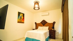 A bedroom with a bed, a nightstand, a lamp, a painting, and an air conditioning unit.