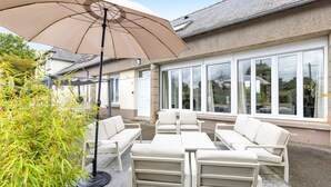 En terrasse med udendørs møbler, en stor parasol og et hus med store vinduer.