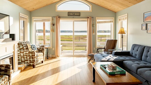 A cozy living room with a wooden ceiling, a large window with a view of a bridge, and a comfortable seating area. - Waterfront Cottage in Stanley Bridge—Bay Views, Sleeps 8, Near Cavendish (North Rustico)