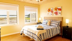A bedroom with a large bed, two windows, a ceiling fan, and artwork on the wall. - Waterfront Cottage in Stanley Bridge—Bay Views, Sleeps 8, Near Cavendish (North Rustico)