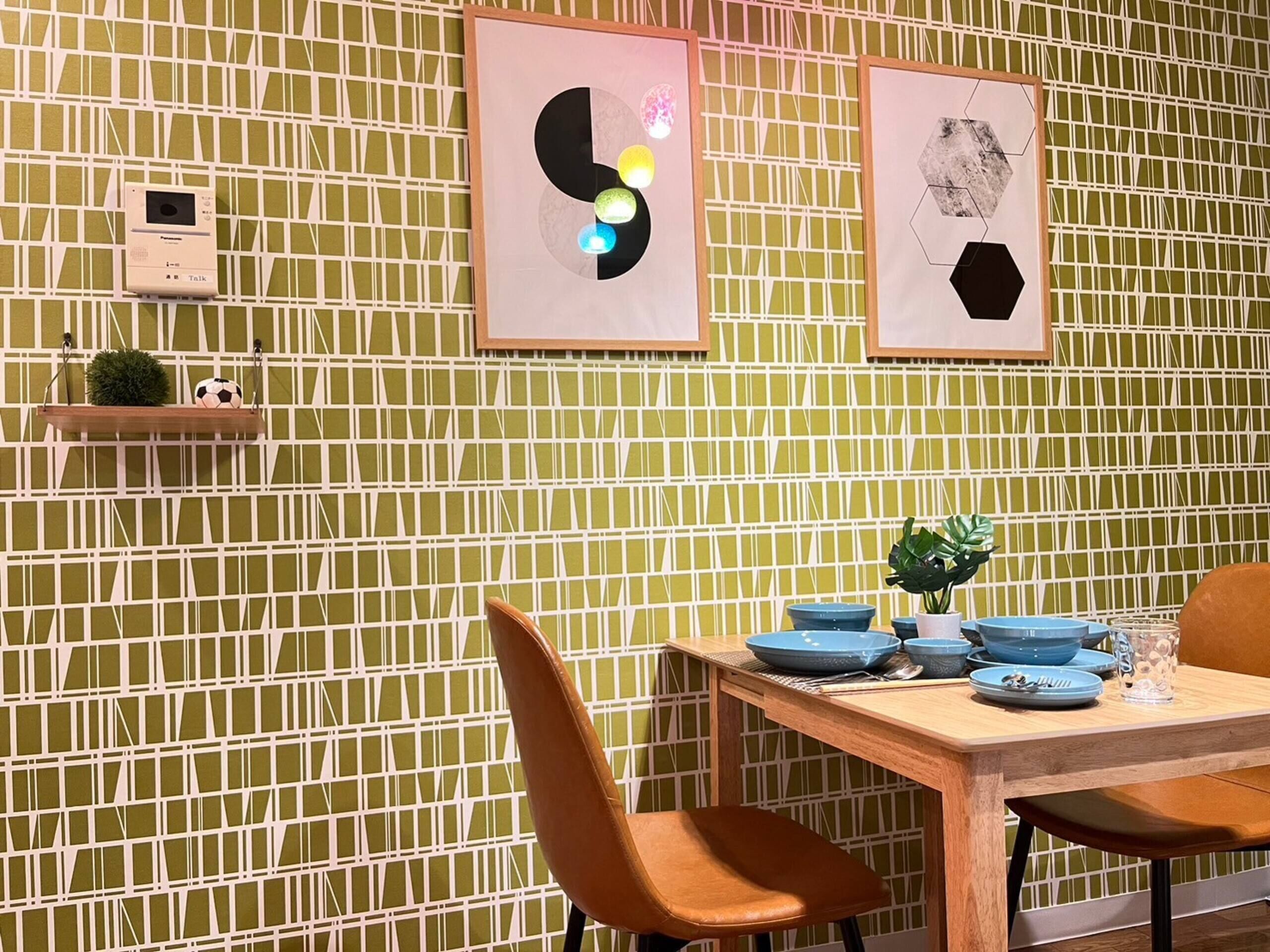 A dining area with a green patterned wall, two framed abstract art pieces, a small shelf with a plant and a soccer ball, and a wooden table with blue plates and a glass pitcher.