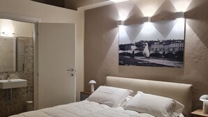 A bedroom with a large bed, a nightstand with a lamp, a wall-mounted picture, and a bathroom with a sink and mirror.