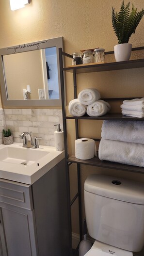 A bathroom with a sink, mirror, towel rack, toilet, and a shelf with toiletries.