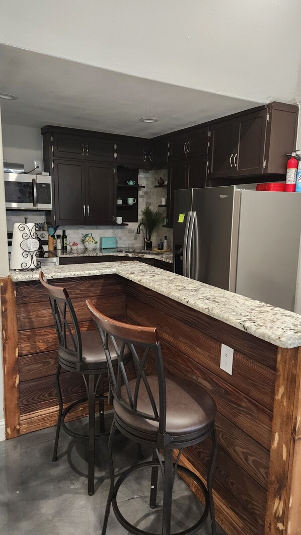 A kitchen with dark wooden cabinets, a stainless steel refrigerator, a microwave, and a granite countertop. There are two bar stools with dark seats.