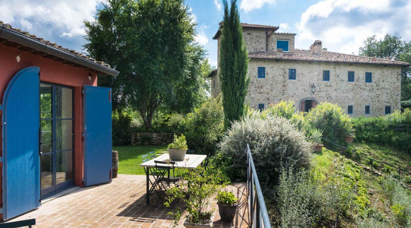 Un bâtiment en pierre avec un toit en tuiles, une porte bleue et un jardin agrémenté d’une table et de chaises.