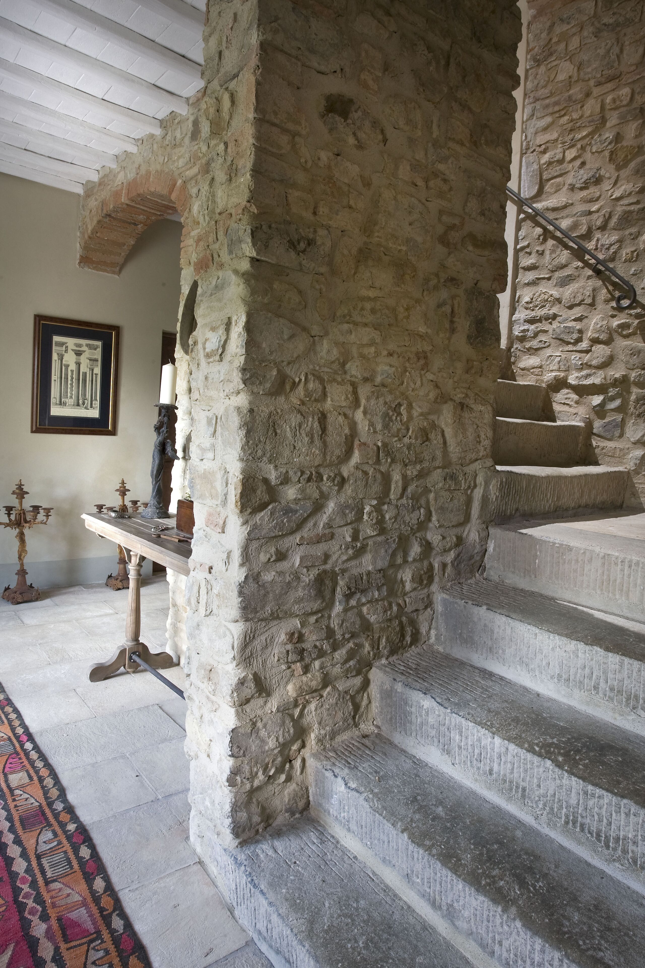 A stone staircase with a handrail, leading to a landing with a framed picture and a candelabra.