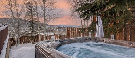 Un jacuzzi rodeado de nieve, árboles y una valla de madera, con vistas a la montaña.
