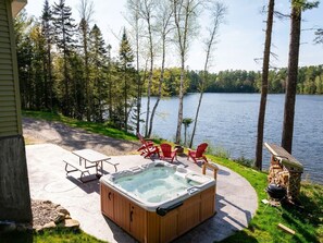 A hot tub with chairs, a table, and a grill by a lake.