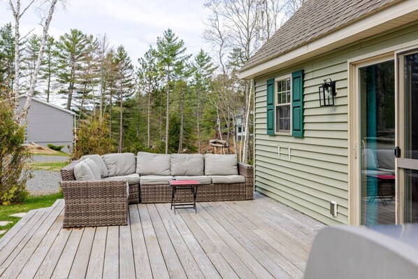 A covered deck with a wicker sofa and a small table, surrounded by a wooden deck and a house with green shutters.
