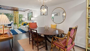 A dining area with a round table, chairs, and a chandelier. There is a mirror on the wall and a window with curtains.