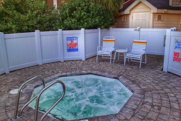 A hot tub with a wooden deck, two lounge chairs, and a white fence.