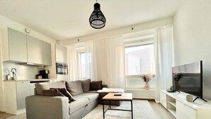 Uma sala de estar moderna com um sofá cinzento, uma mesa de centro, uma televisão e uma área de cozinha com armários e eletrodomésticos.