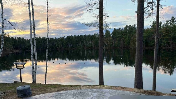 Un patio de concreto con una silla Adirondack roja, un farol y vista a un lago tranquilo rodeado de árboles.