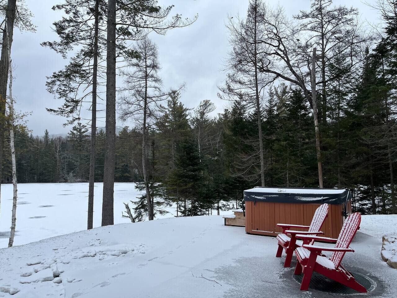 A wooden hot tub with two red Adirondack chairs in a snowy forest setting.