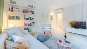A modern living room with a white sofa, a red chair, a bookshelf, a TV, and a small table with drinks and fruit.