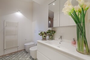 A modern bathroom with a white sink, mirror, and toilet. There are two glass vases with flowers on the sink counter.