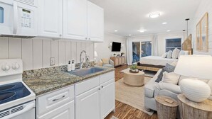 Eine moderne Küche mit weißen Schränken, einer eingebauten Mikrowelle und einem Herd. Es gibt eine Essecke mit einem runden Tisch und einer Couch. Der Raum verfügt über ein großes Fenster und ein Bett mit Blick.