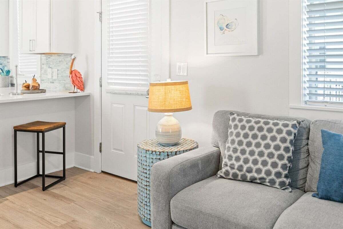 Ein modernes Wohnzimmer mit einem grauen Sofa, einem kleinen Holzschemel, einem runden Beistelltisch mit Lampe und einem dekorativen Vogel auf der Ablagefläche.