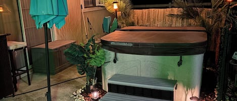 A hot tub with steps, surrounded by potted plants and a patio area with a bench and umbrella.
