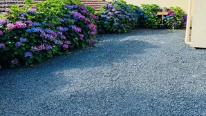 A gravel driveway leading to a house with a tiled roof and a chimney. There are flowering bushes along the driveway.