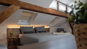 Une chambre avec un plafond de style industriel, un lit avec une literie grise, une petite table de chevet avec une plante et un bureau avec une lampe.