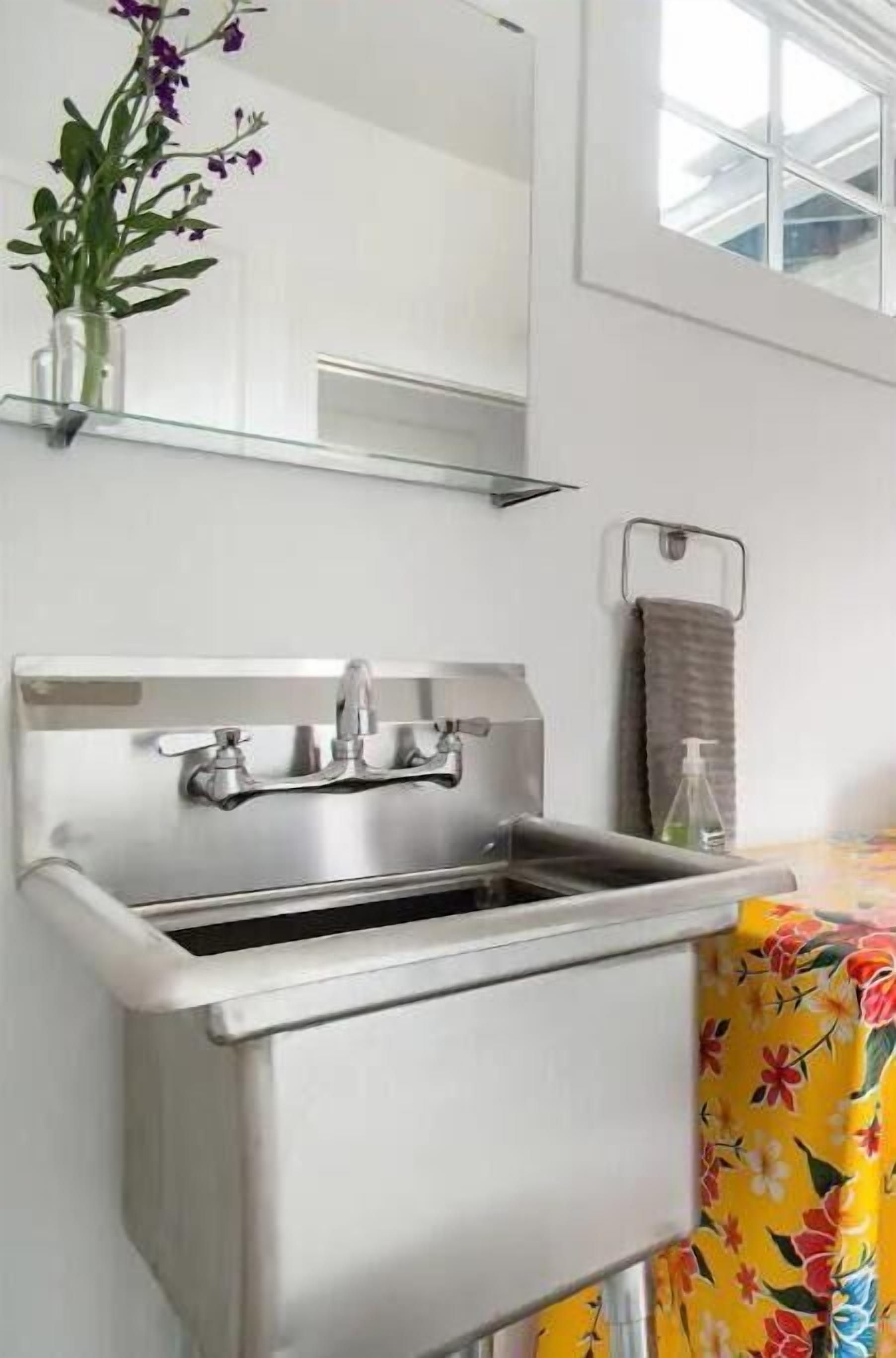 A bathroom with a stainless steel sink, a mirror above it, a towel rack, and a vase with purple flowers on the wall.