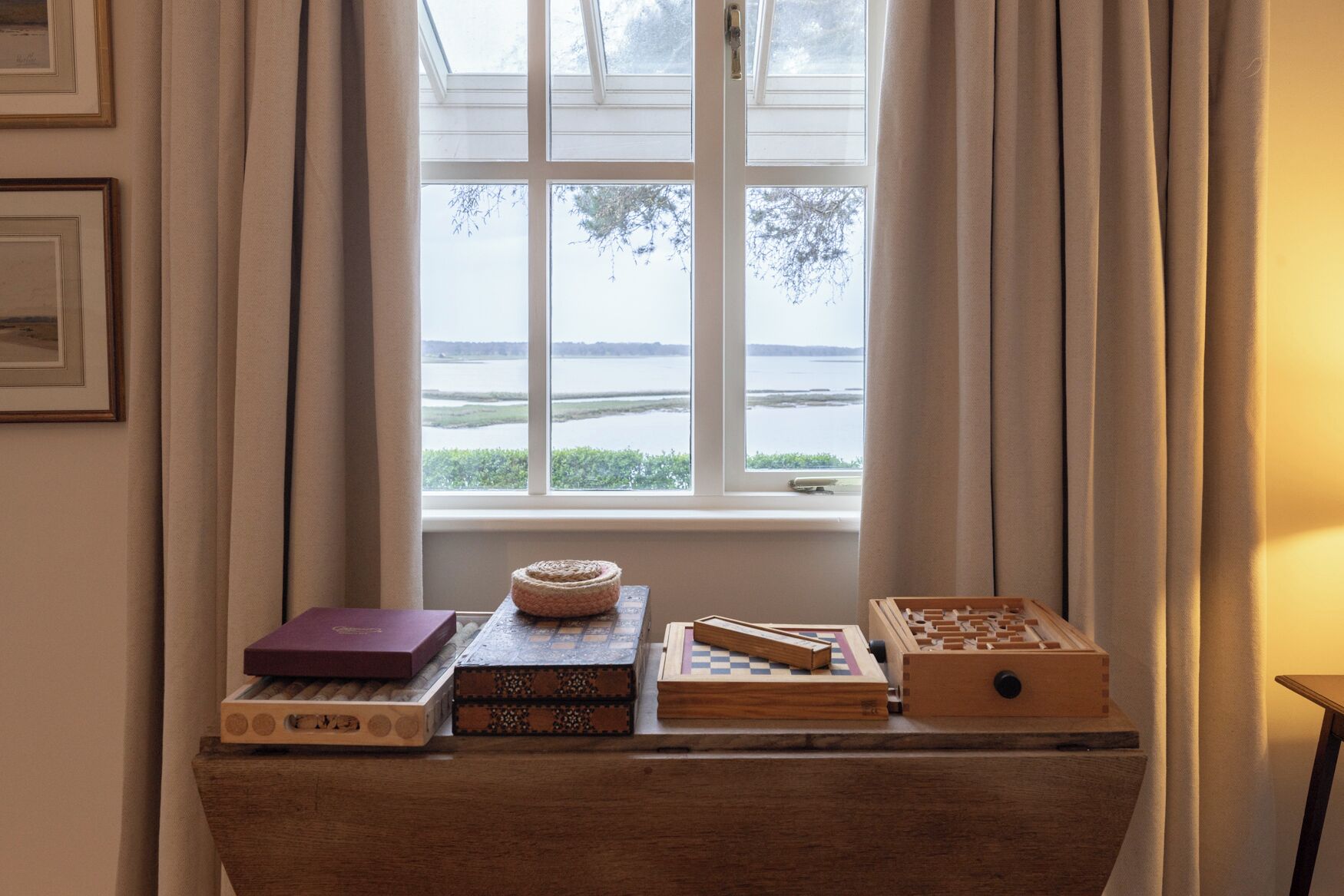 Une table en bois, sur laquelle sont posés des livres et des jeux de société, se trouve près d’une fenêtre donnant sur un lac.