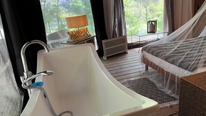A bedroom with a large bathtub, a bed with a canopy, a window with curtains, and a lamp on a side table. - Prehistoric Lodge (Vallon-Pont-d'Arc)