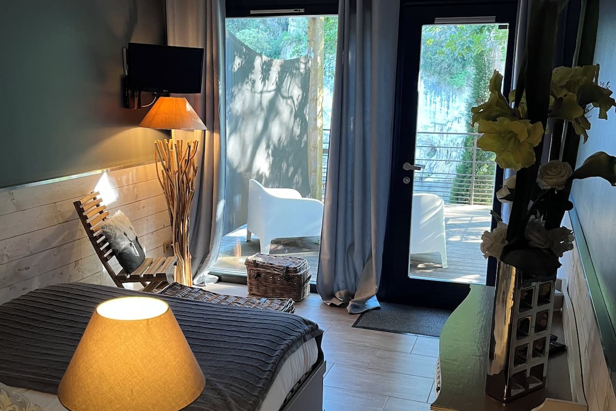 A bedroom with a bed, a bedside table with a lamp, a wooden side table, and a view of the outdoors through a window.