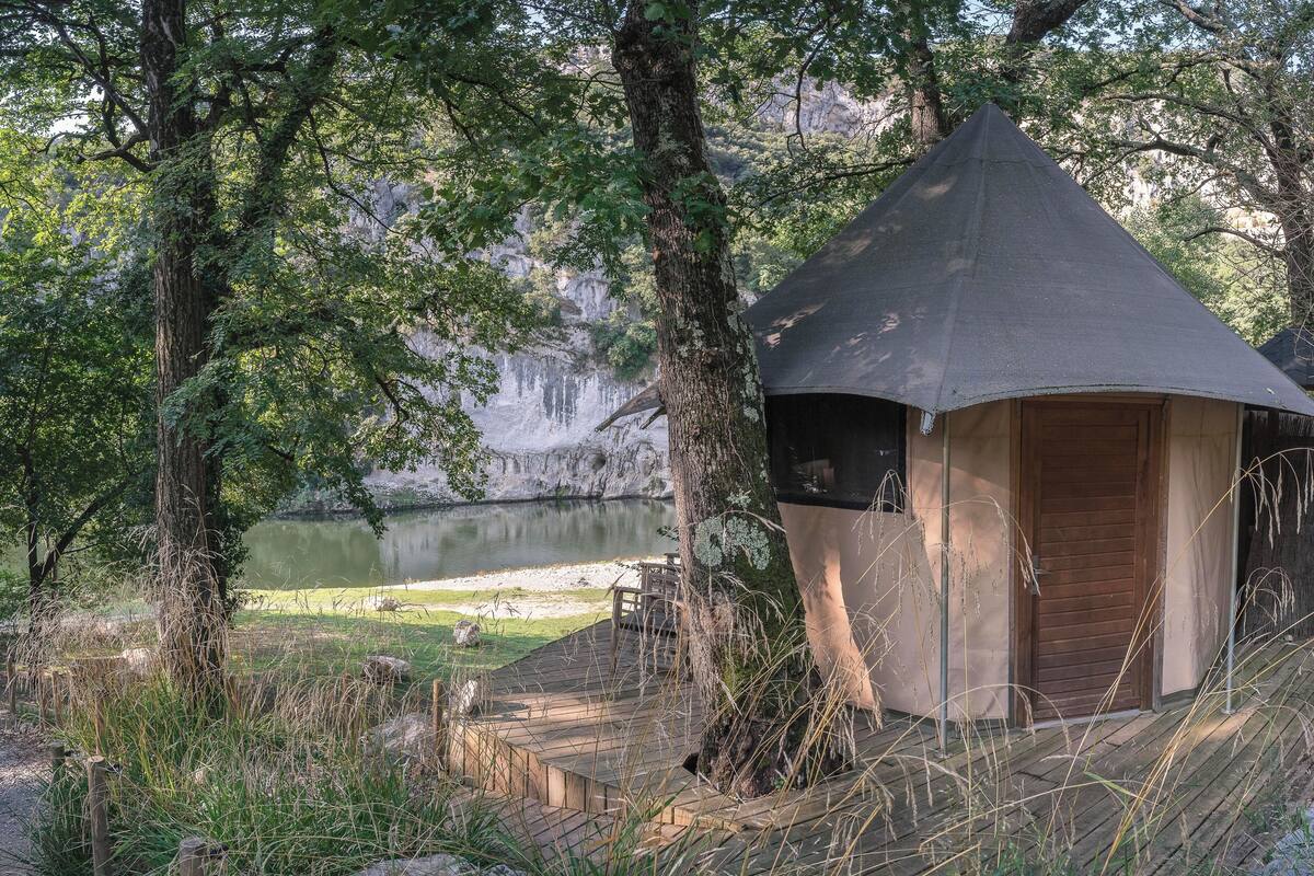 A wooden cabin with a conical roof, surrounded by trees and a lake.