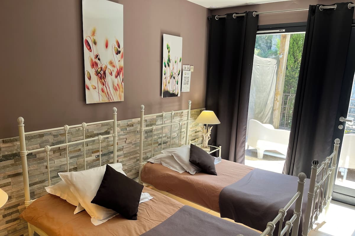 A hotel room with two beds, a stone wall, artwork, a lamp, and a view of greenery.