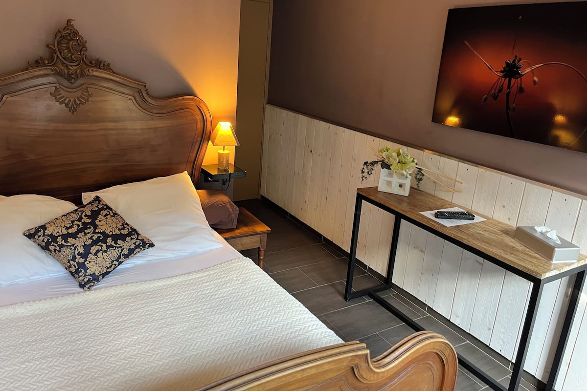 A neatly made bed with a patterned pillow, a bedside lamp, a small table with a vase of flowers, and a painting on the wall.