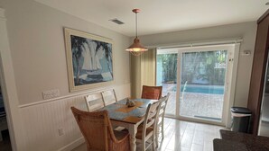 Un comedor con una mesa dispuesta para seis personas, rodeada de sillas de mimbre, un cuadro sobre la mesa y vistas a una piscina a través de puertas de vidrio.