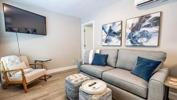 A modern living room with a grey sofa, a wooden chair, and two ottomans. A flat-screen TV is mounted on the wall, and there are abstract paintings above the sofa. - Ocean Breezes (Lantana)