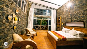 A room with a bed, a chair, a small table, a bookshelf, and a large window with curtains. - Imbogo Diners and Lounge (Kisoro)