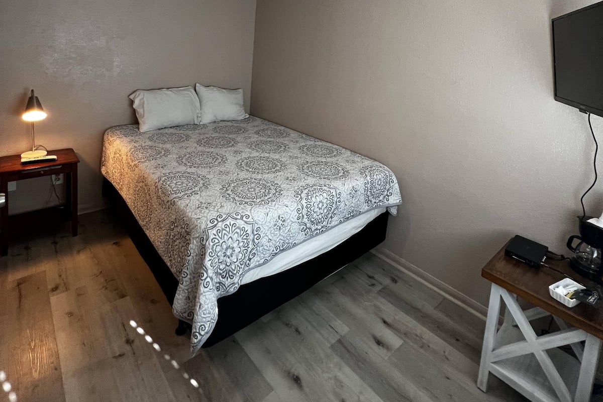 Un dormitorio con cama, una mesita de noche con lámpara, un televisor montado en la pared y piso de madera.