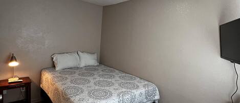 A bedroom with a bed, a bedside table with a lamp, a TV mounted on the wall, and a wooden floor.
