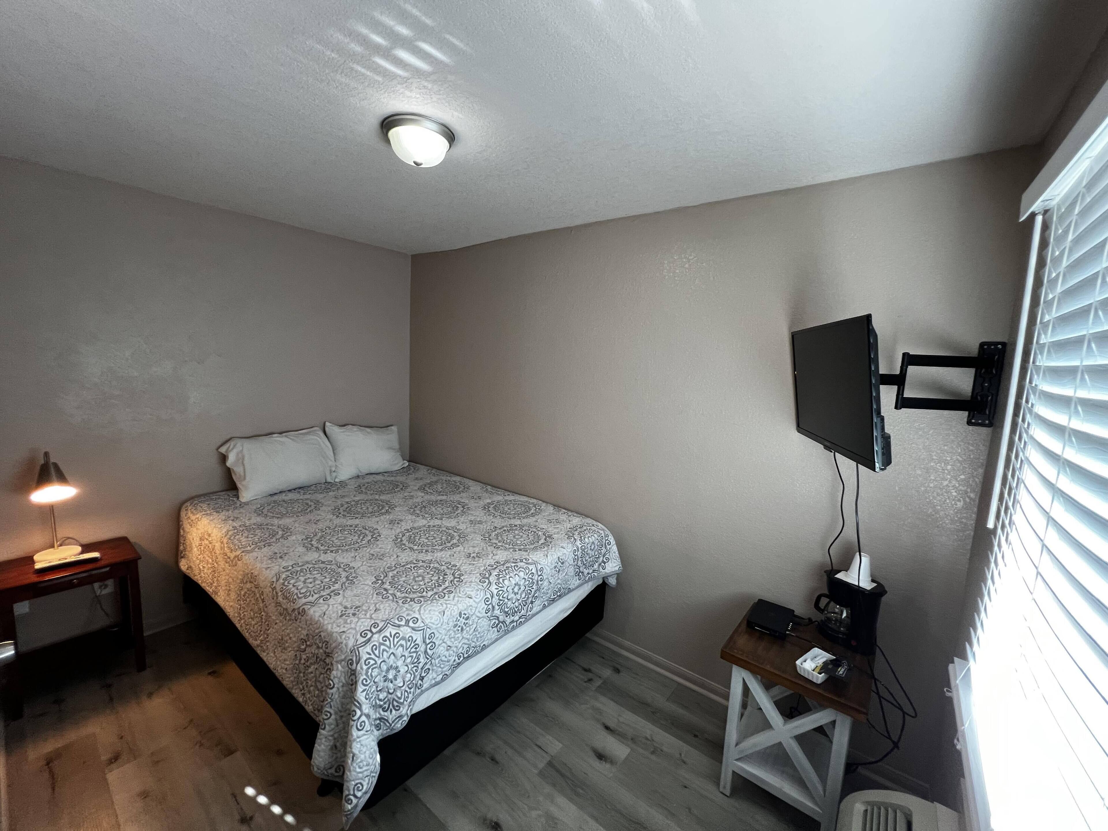 A bedroom with a bed, a nightstand, a desk, a television, and a window with blinds.