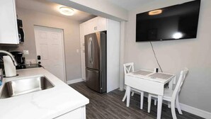 A compact kitchen with a stainless steel refrigerator, a white dining table with chairs, and a flat-screen TV mounted on the wall. - Coastal Breezes -Walk to the Beach (Lantana)