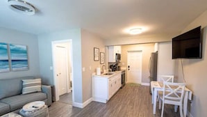 A modern living room with a gray sofa, a dining area with white chairs, a kitchen with white cabinets, and a flat-screen TV mounted on the wall. - Coastal Breezes -Walk to the Beach (Lantana)