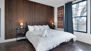 A modern bedroom with a large bed, wooden panel wall, two bedside tables with lamps, and a window with curtains.