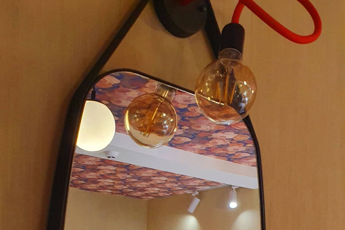 A mirror with a wooden frame reflecting a bed, pillows, and a wall-mounted light fixture.