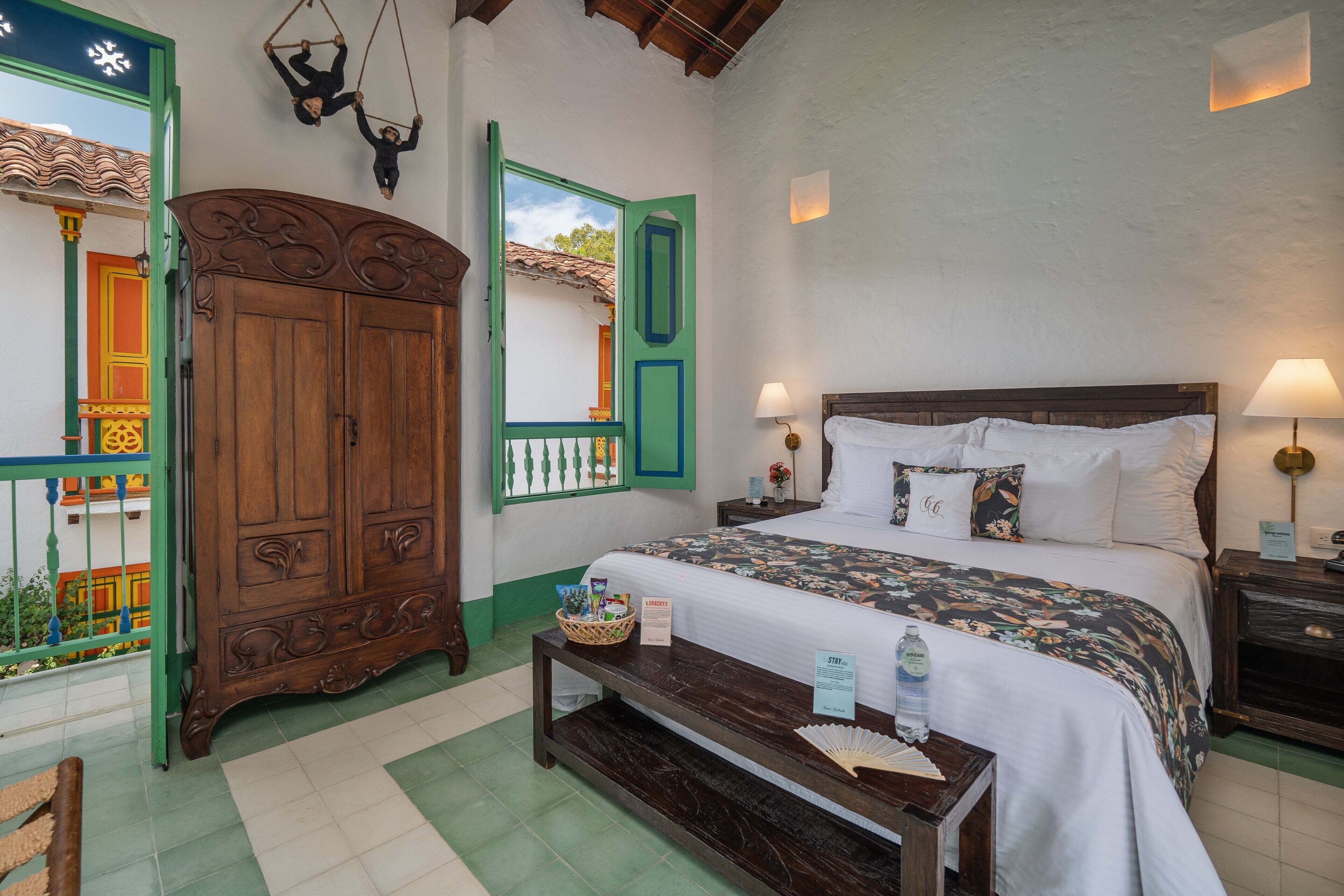 A bedroom with a bed, bedside tables, a wooden wardrobe, and a window with a view of a colorful building.