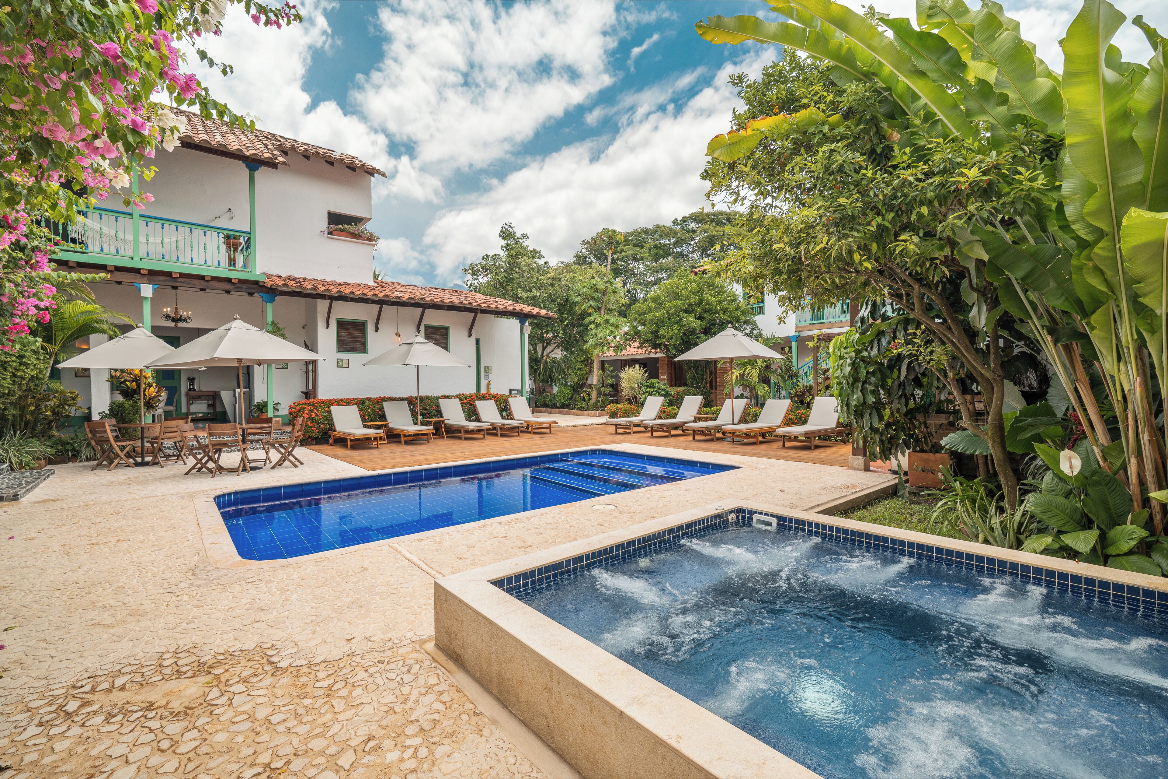Zona de piscina con sillones de descanso y sombrillas, rodeada de exuberante vegetación y un edificio blanco con tejado de tejas.