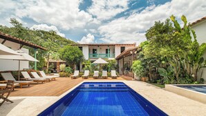 Una piscina con sillones de descanso y sombrillas, rodeada de exuberante follaje y un edificio con balcón.