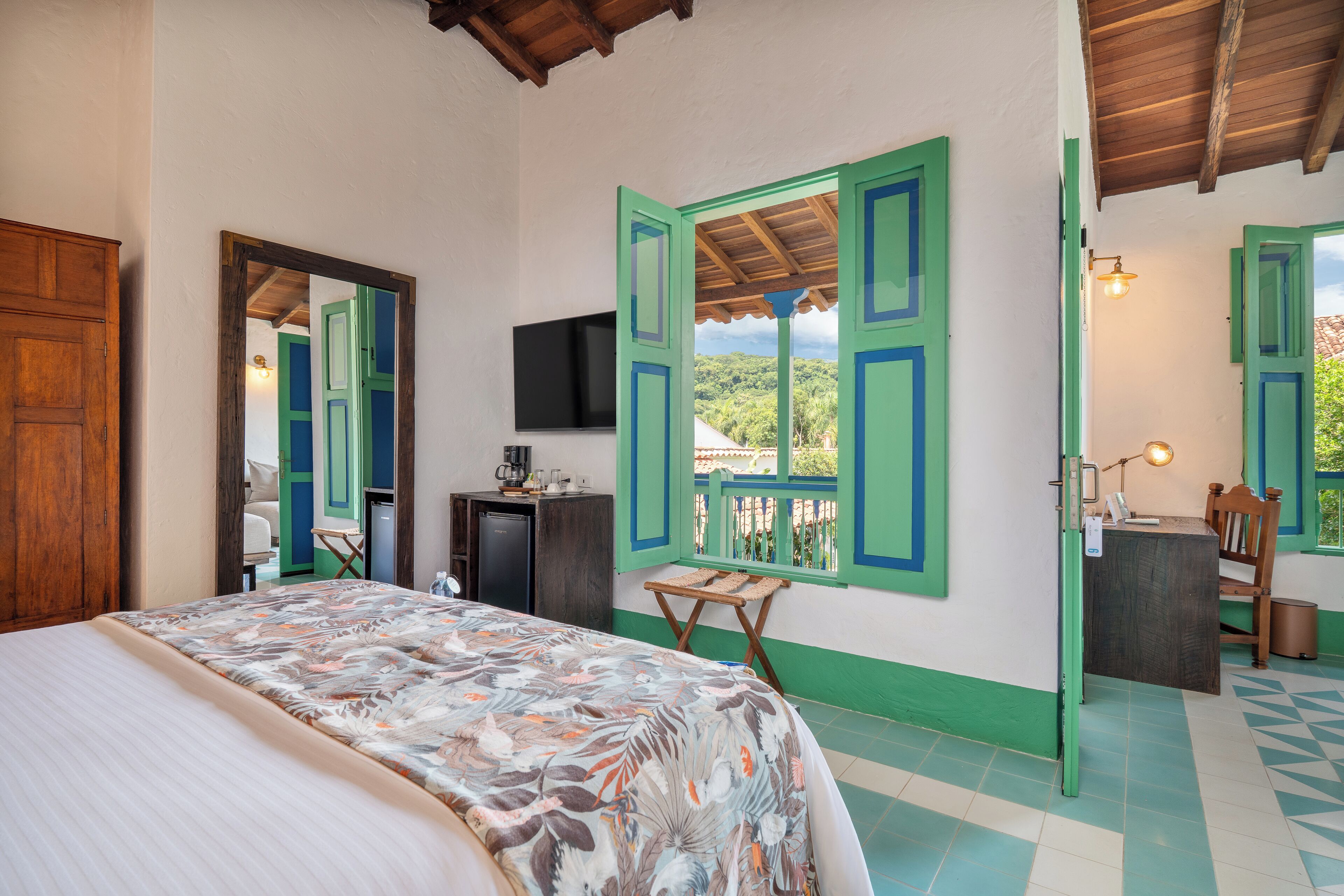 Ein Schlafzimmer mit einem Bett, einem Schreibtisch, einem Sessel, einem Fernseher und einem Balkon mit grünen Fensterläden.